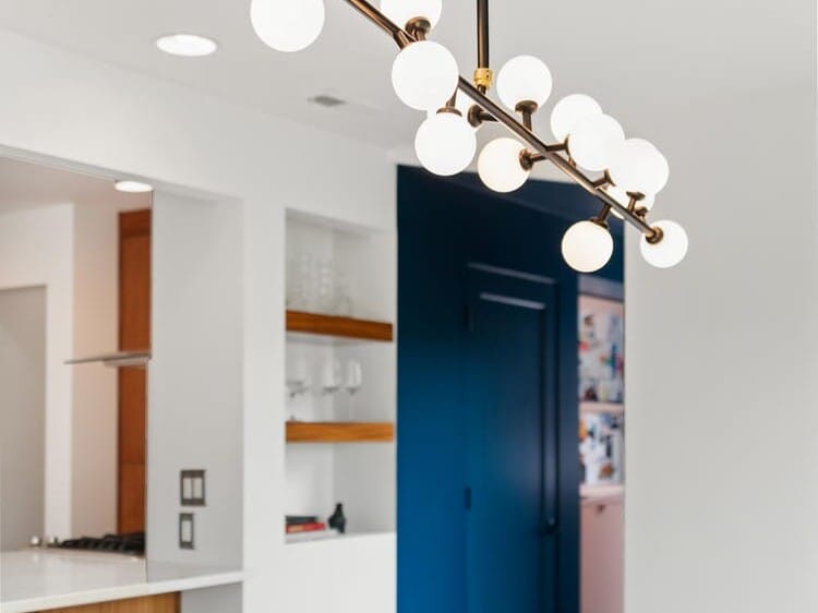 Modern dining room with a sleek black table and a contemporary chandelier featuring spherical white lights. A deep blue accent wall and open shelves with decor are visible in the background, along with a doorway leading to the kitchen.