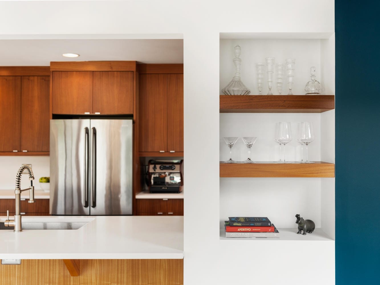 Modern kitchen with stainless steel appliances, wooden cabinets, and a white countertop with a sink. Shelves nearby display glassware, books, and a small decorative hippo.