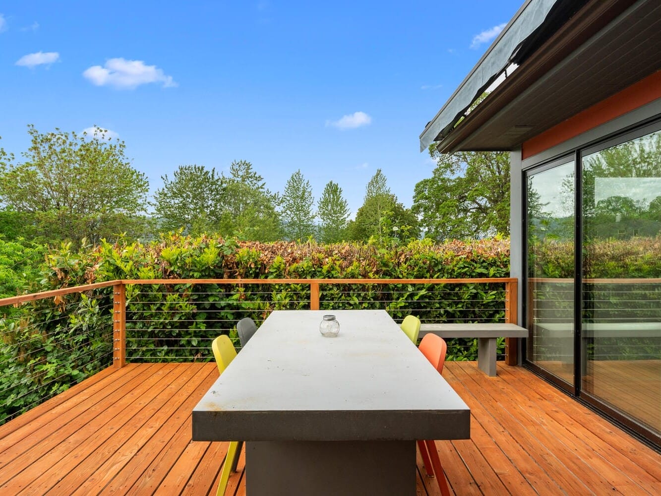 A modern wooden deck with a large concrete table surrounded by colorful chairs. The deck overlooks lush green trees and bushes under a clear blue sky. Glass doors on the right reflect the outdoor scene.