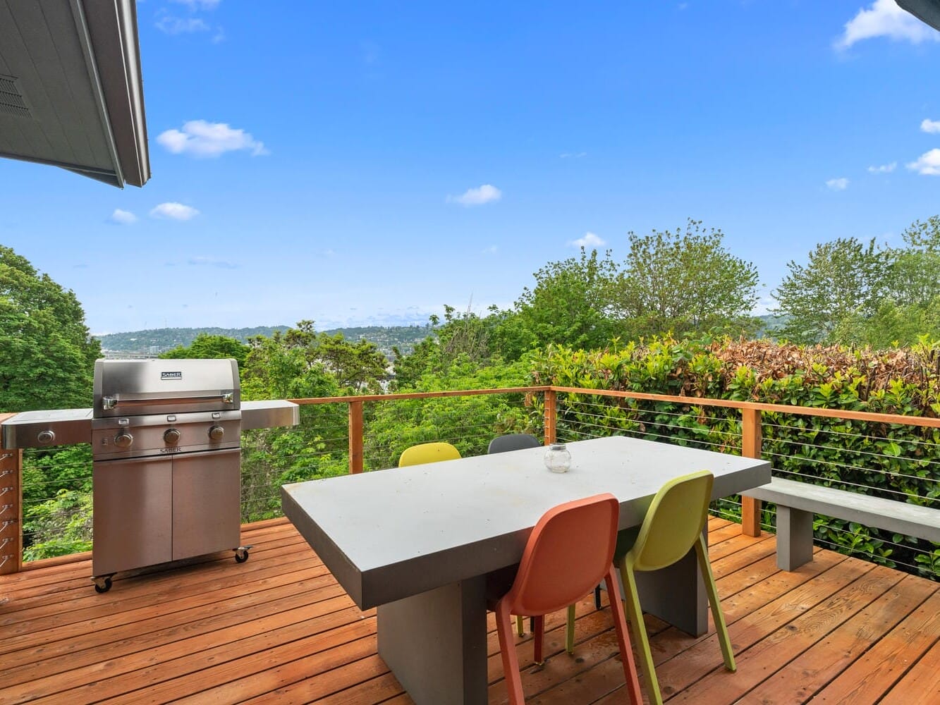 Outdoor patio with a modern grill and a concrete table surrounded by colorful chairs, set on a wooden deck. Lush green trees and bushes provide a natural backdrop, with a distant view of water and hills under a blue sky.