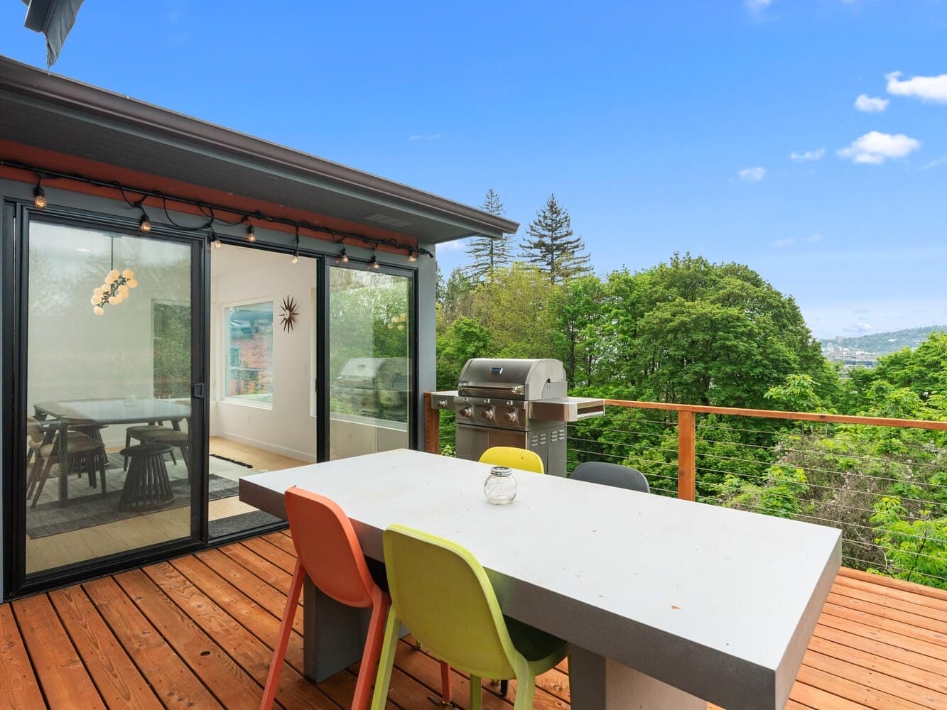 A patio with a white dining table and colorful chairs overlooks a lush green landscape. A barbecue grill is nearby. Sliding glass doors lead into a modern dining room. The sky is clear and blue.