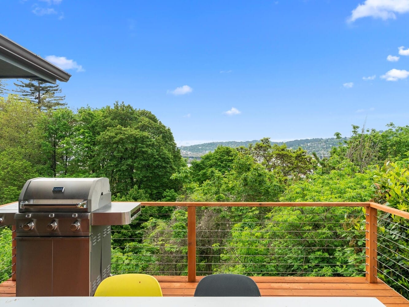 A balcony with a stainless steel grill overlooks a lush green landscape and distant hills under a clear blue sky. The railing is wood with cable wiring, and there are colorful chairs visible in the foreground.