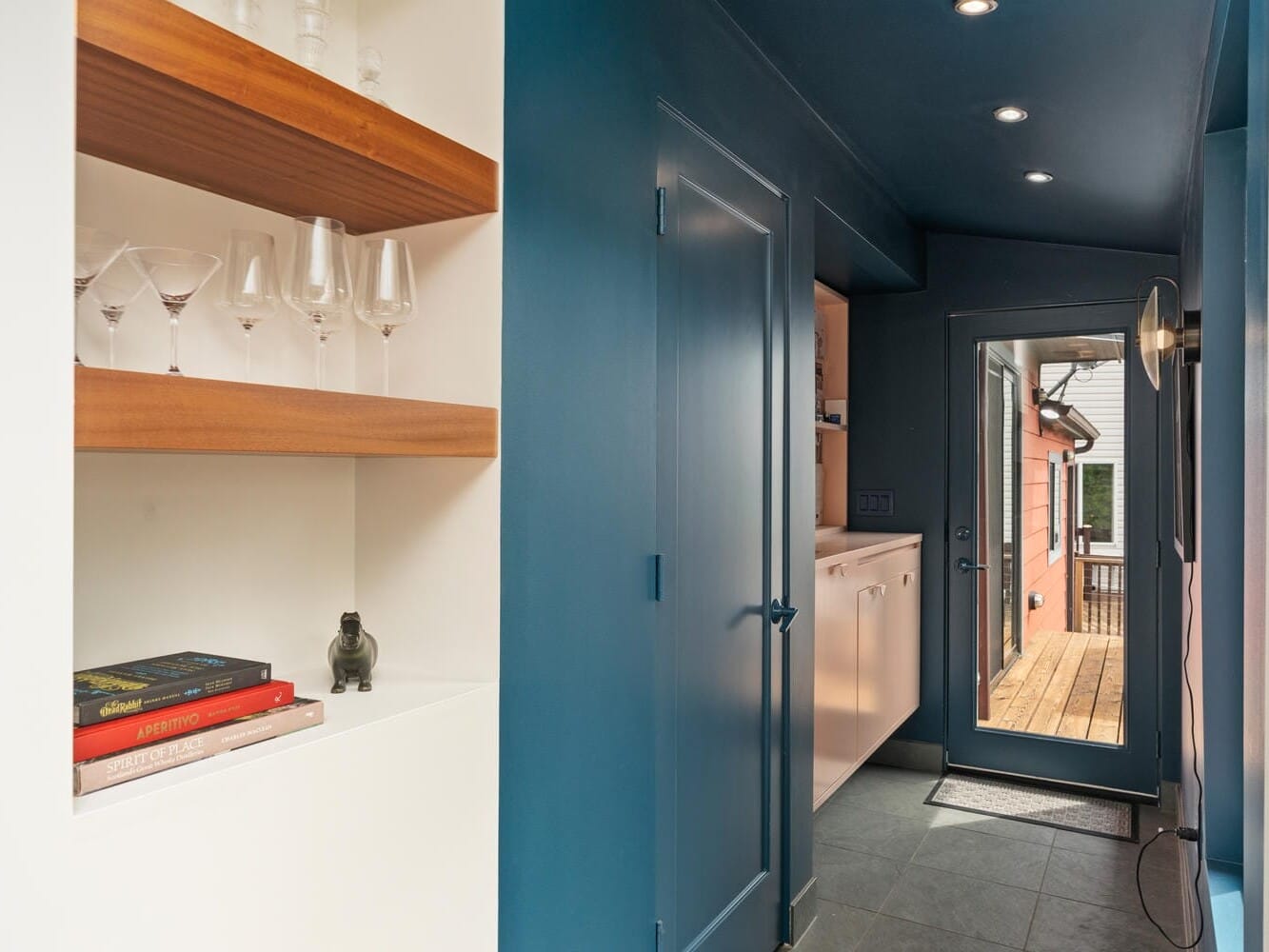 A narrow hallway with dark blue walls and ceiling. Shelves on the left hold glassware, books, and a small sculpture. A door on the right opens to a wooden deck, leading outside. Recessed lighting illuminates the space.