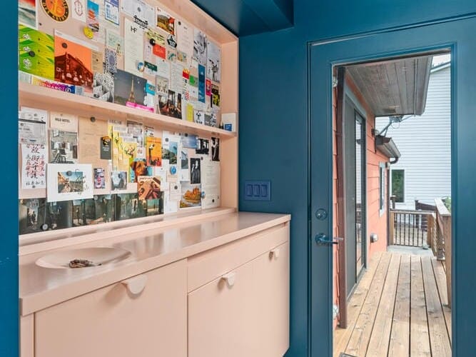 A mudroom with blue walls features a pale pink cabinet and sink. The wall above the sink hosts a large collection of colorful postcards and photos. A glass door leads to a wooden deck outside. A small mat is placed on the floor in front of the door.