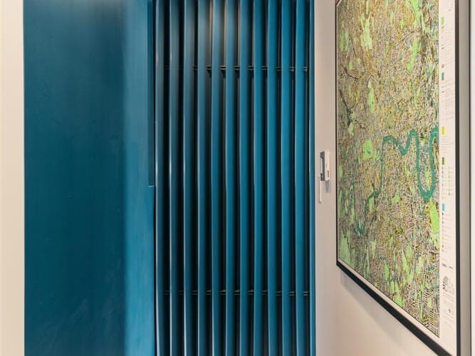 A narrow hallway with blue walls and wood flooring leads to a patterned dark blue panel. A colorful map is framed and hung on the right wall. A ceiling light illuminates the space, creating a modern and minimalist atmosphere.