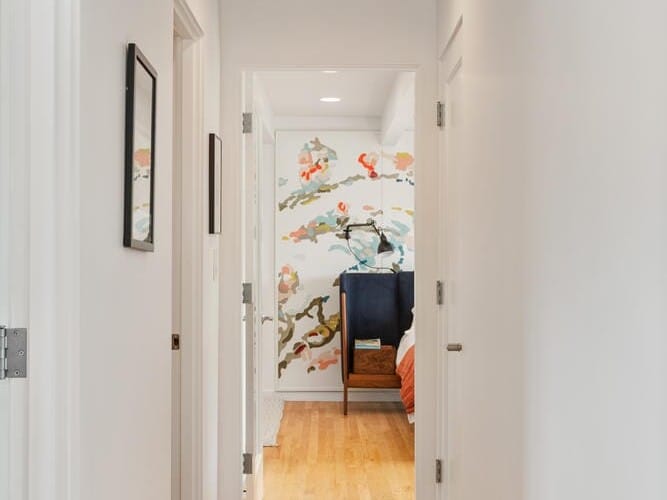 A hallway with wooden floors and white walls leads to a bedroom. The ceiling is painted blue. The bedroom at the end has a colorful wall mural and minimal furniture visible. Doors to additional rooms line the hallway on both sides.