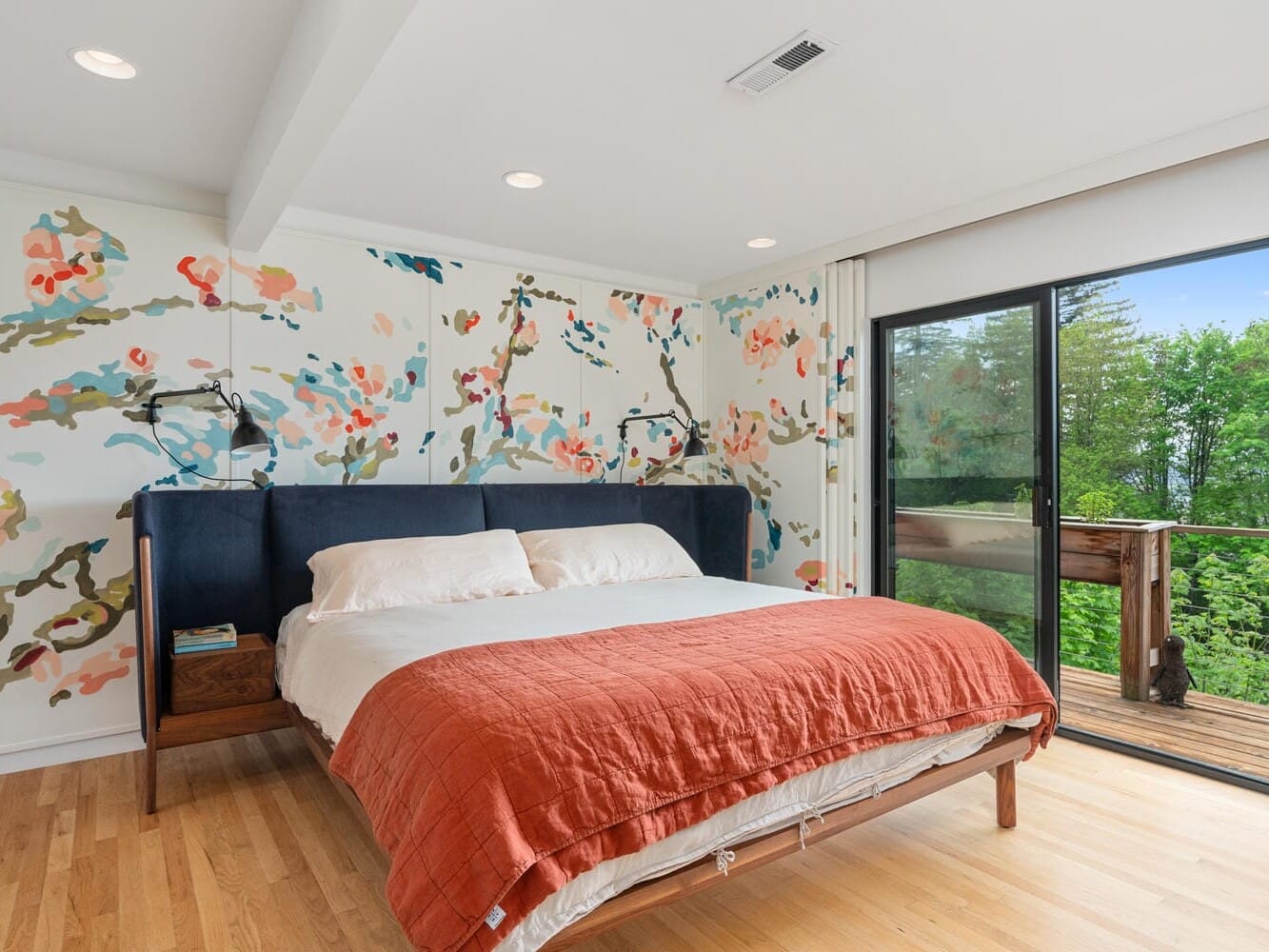 A modern bedroom with a large bed featuring a rust-colored blanket and white pillows. The wall behind the bed is decorated with a colorful floral mural. Glass sliding doors on the right lead to a wooden deck with greenery outside. Wood flooring is visible.