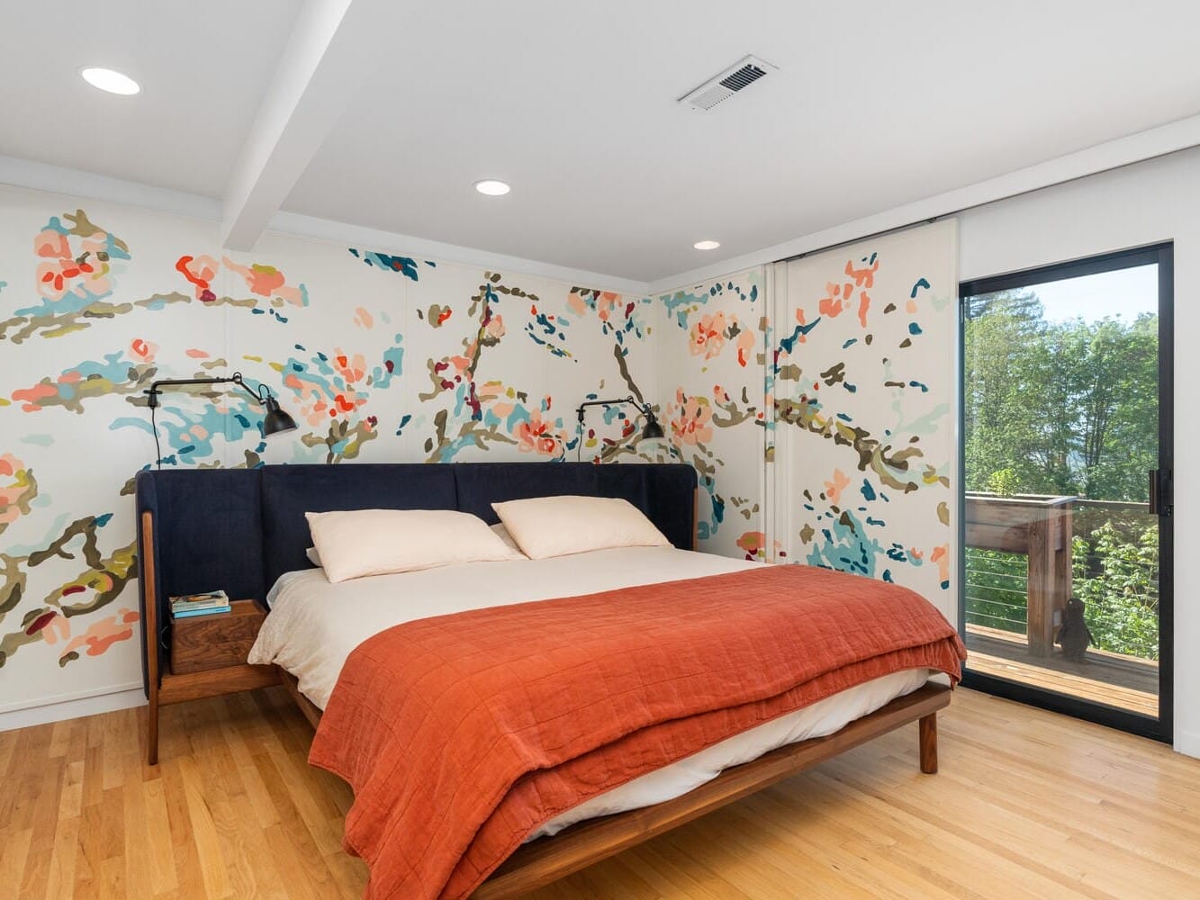 A modern bedroom with a king-size bed featuring a dark blue headboard and an orange quilt. The walls have a colorful floral mural. Theres a sliding glass door on the right leading to a balcony with a view of trees. The floor is light wood.