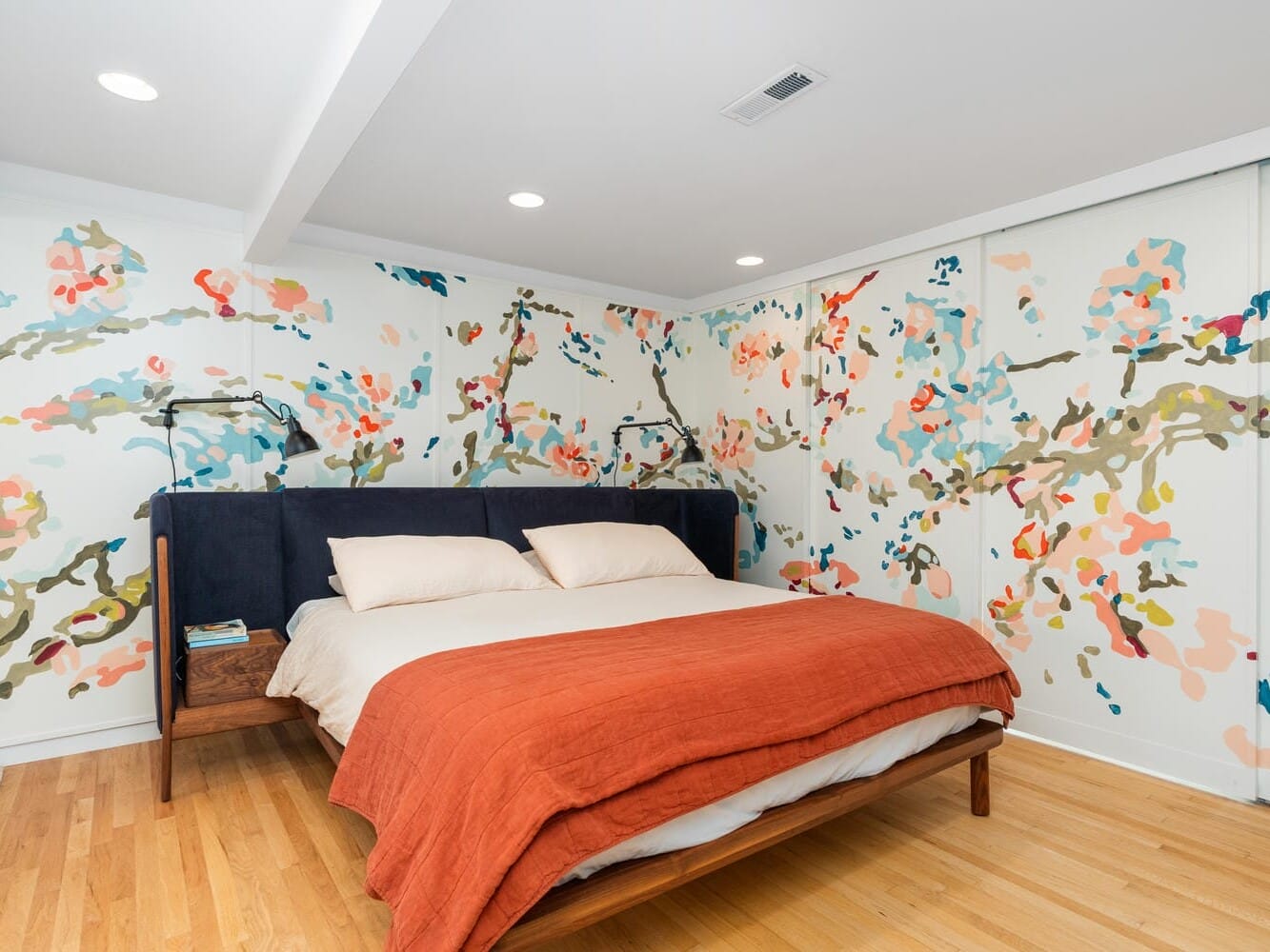 A cozy bedroom features a bed with a dark headboard, light bedding, and a rust-colored blanket. The walls are adorned with vibrant floral wallpaper, and there is wood flooring, creating a warm and inviting atmosphere.