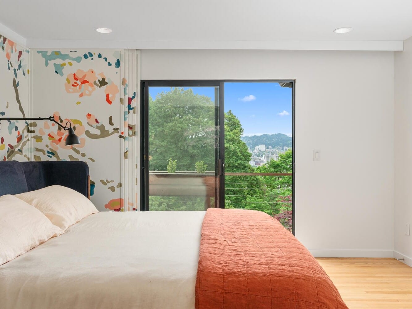 A bedroom with a large bed featuring beige and orange bedding. Theres a colorful floral accent wall and a sliding glass door leading to a balcony. Outside, there is a view of lush green trees and a distant hill.