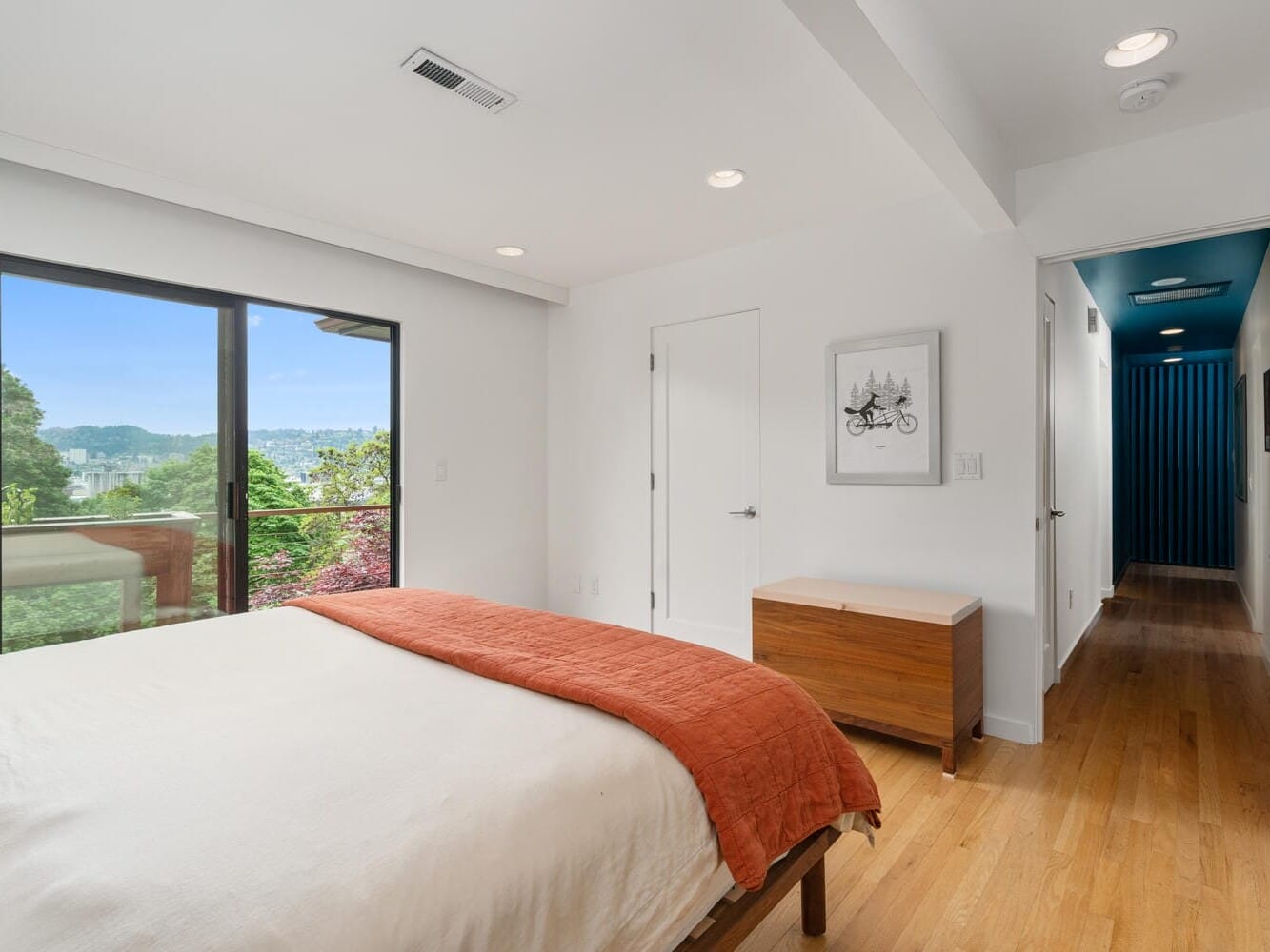 A bright bedroom with a queen-sized bed covered in white and orange bedding. A large window offers a view of greenery and hills. Theres a wooden chest at the foot of the bed, and a hallway with a blue accent wall is visible.