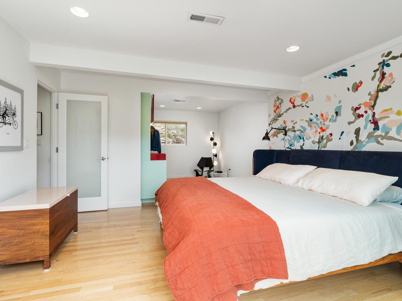 Modern bedroom with a large bed featuring a colorful abstract mural on the wall behind it. The bedding is white with a red-orange blanket. A wooden dresser is on the left, and theres natural light coming in from windows.