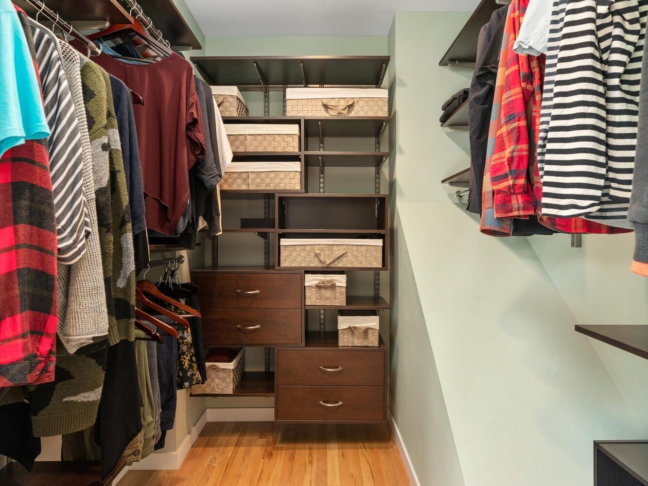 A neatly organized walk-in closet with shelves and hanging space. Clothes, including shirts and jackets, hang on the right, while folded items and storage baskets are on the left. The floor is wooden, and the walls are light green.