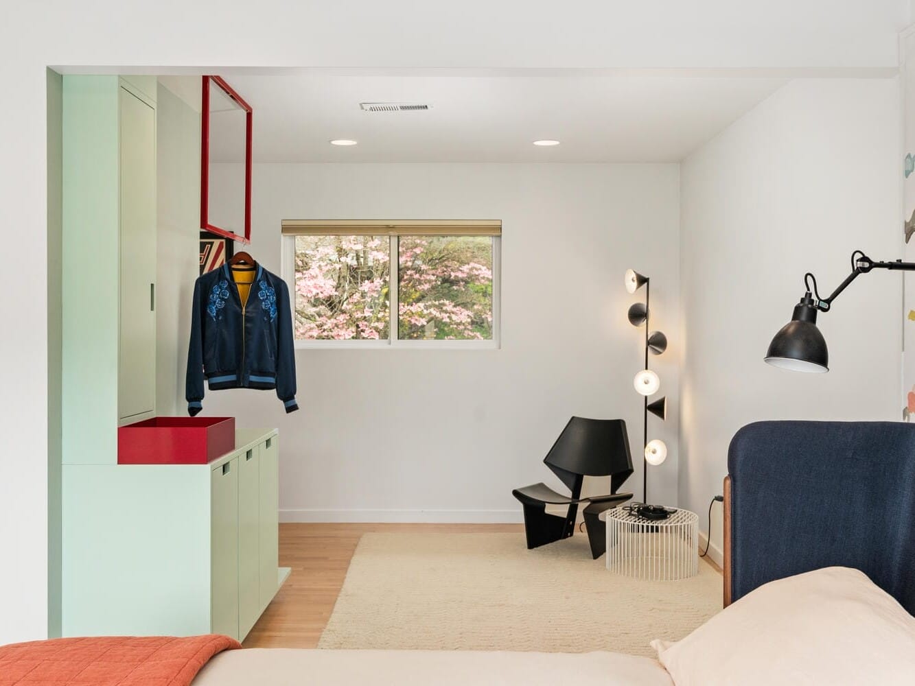 A bright room with a mint green cabinet and red countertop. A jacket hangs on an open red frame. Theres a window with a view of trees, a modern light fixture, black chair, and small table. A bed with beige and orange bedding is partially visible.