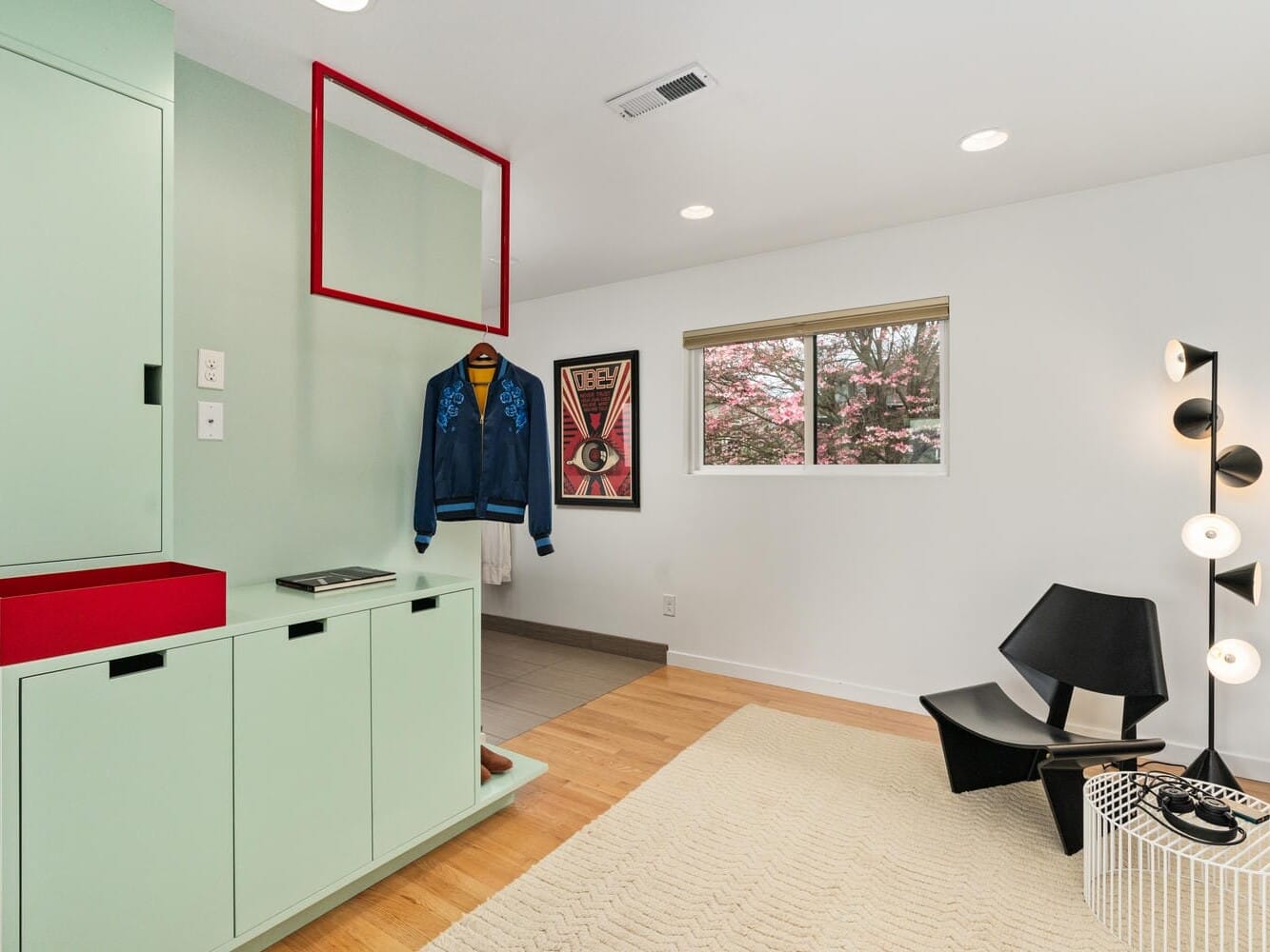 A modern room with mint green cabinets and a red-framed mirror on the wall. Theres a black chair, a multi-bulb floor lamp, and a framed artwork. The window shows a view of a tree with pink blossoms. Wooden floor and tan rug complete the scene.