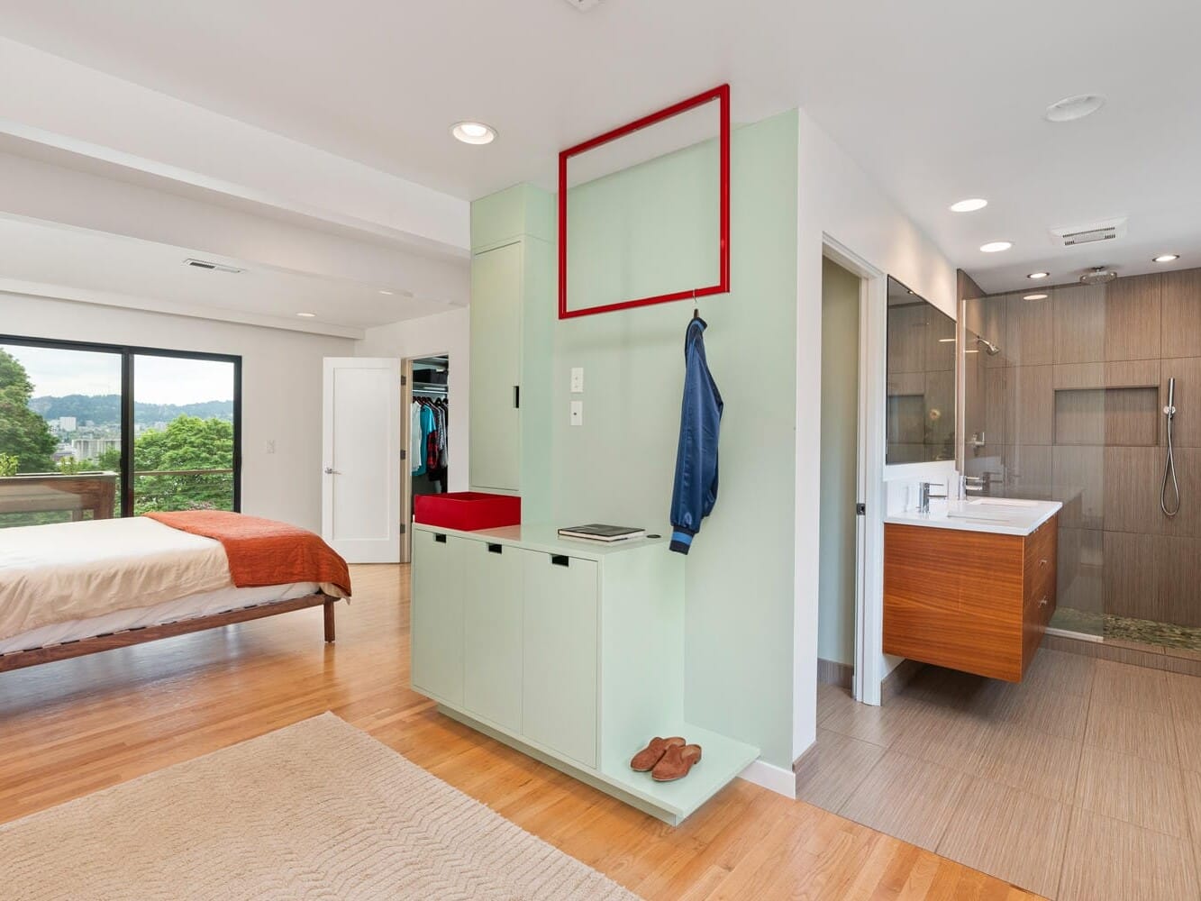 Modern bedroom with a built-in open bathroom. Features a bed with orange bedding, a green wall with hooks and a red accent shelf, and wooden flooring. Glass doors open to a balcony with a view of greenery.