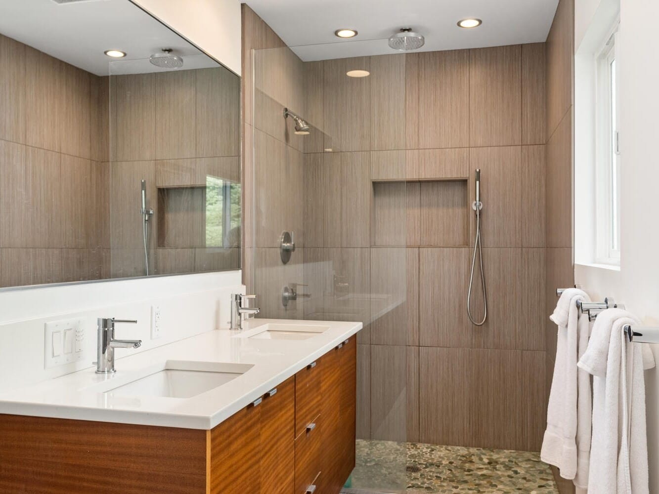 Modern bathroom with a large mirror above a double sink vanity with wooden drawers. Theres a glass-enclosed shower with a rainfall showerhead and tiled walls. White towels hang on a rail by the window, and pebble stone flooring is visible in the shower.