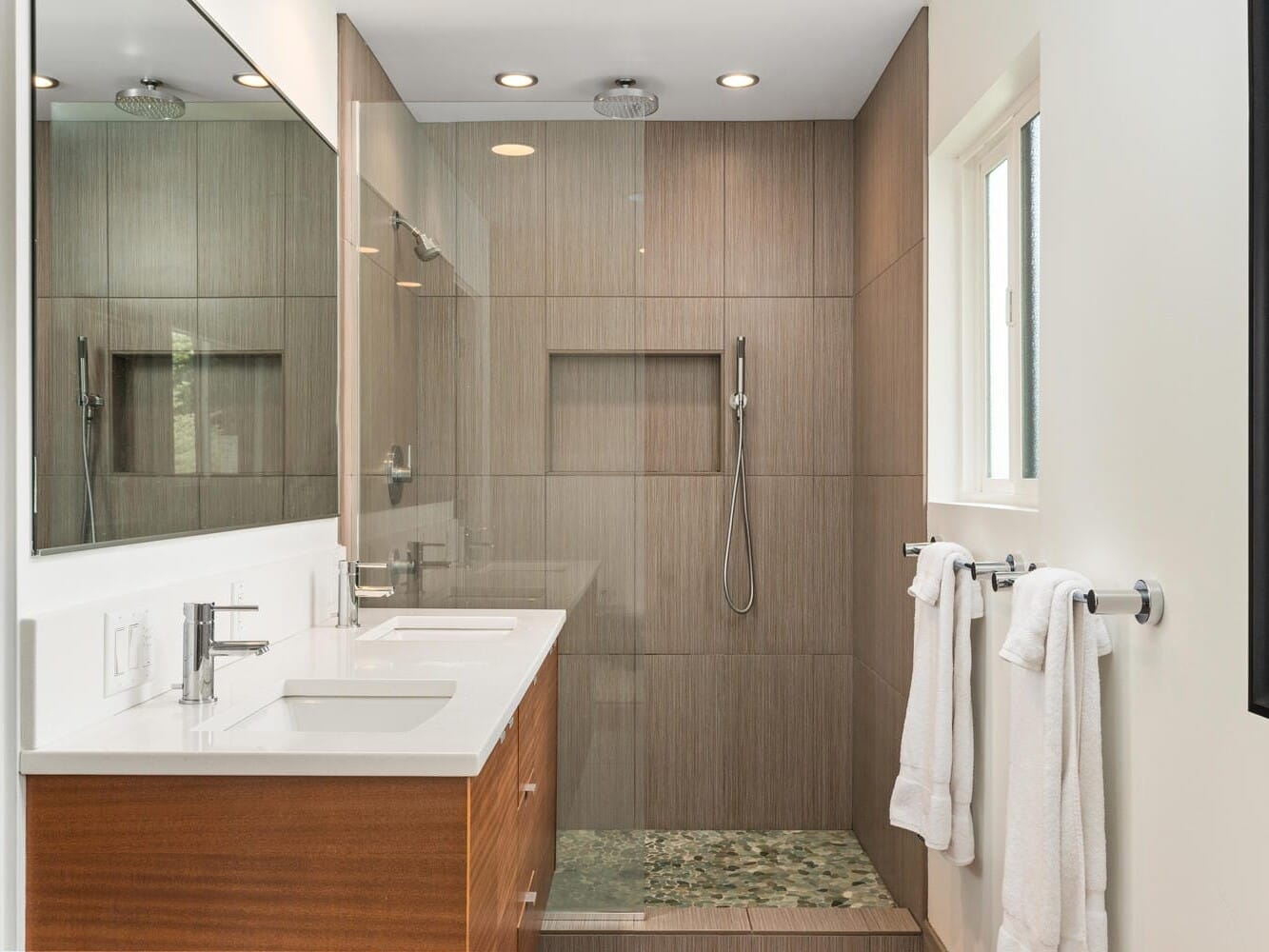 Modern bathroom featuring a wooden vanity with double sinks, a large mirror, and a walk-in shower with a rain showerhead. White towels hang on a rack, and a red and black art piece decorates the wall. Natural light enters from a window.