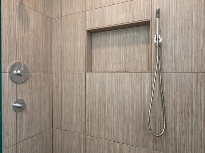 A modern shower with vertical beige tiles and a pebble stone floor. It features a wall-mounted overhead showerhead, a handheld shower on a rail, and a built-in niche for storage. The overall design is sleek and minimalistic.