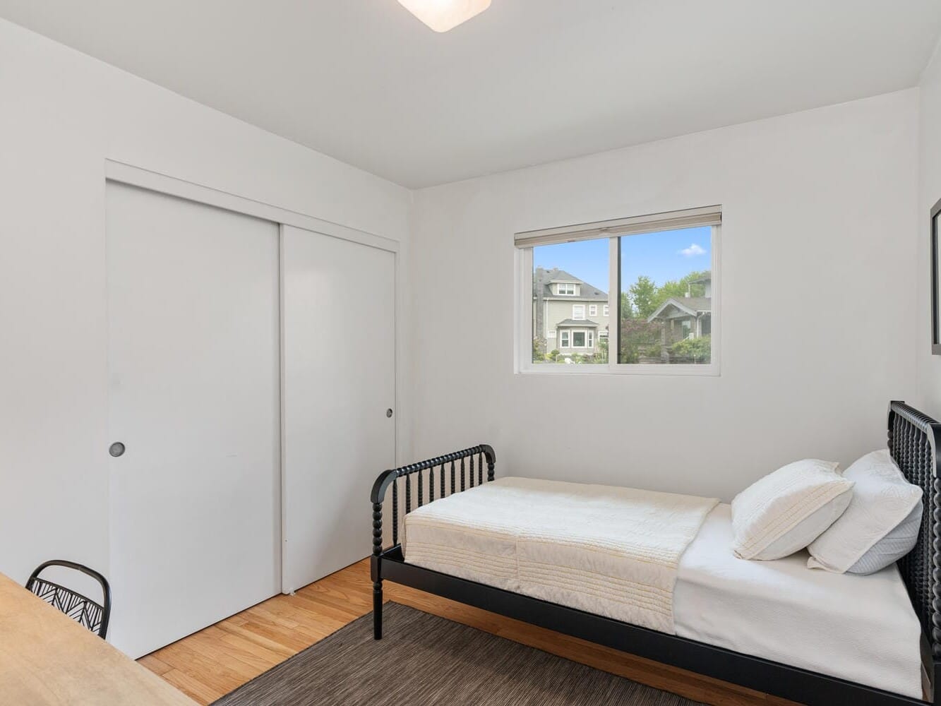 A minimalist bedroom with a single bed, white walls, and a window showing a view of houses and greenery outside. Theres a wooden floor, a wooden desk with a chair to the left, and a sliding closet door on the left wall.