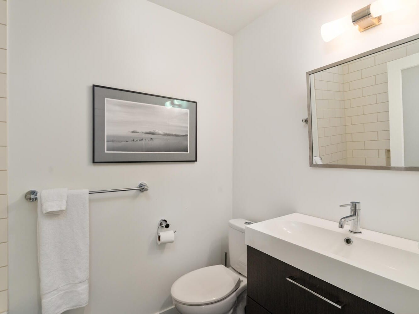 A modern bathroom with a white sink, silver faucet, and dark vanity. A large mirror hangs above the sink. Theres a wall-mounted towel bar with a white towel, and a framed black-and-white photo on the wall. A toilet and toilet paper holder are visible.