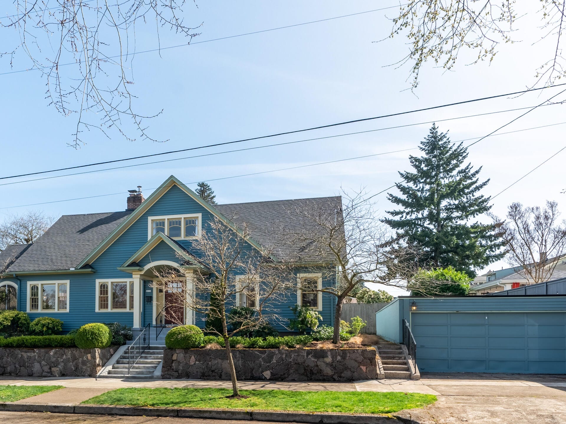 A charming blue house with a steep roof, a stone foundation, and a neatly trimmed hedge in front. Theres a two-car garage on the right, and leafless trees frame the scene under a clear blue sky.