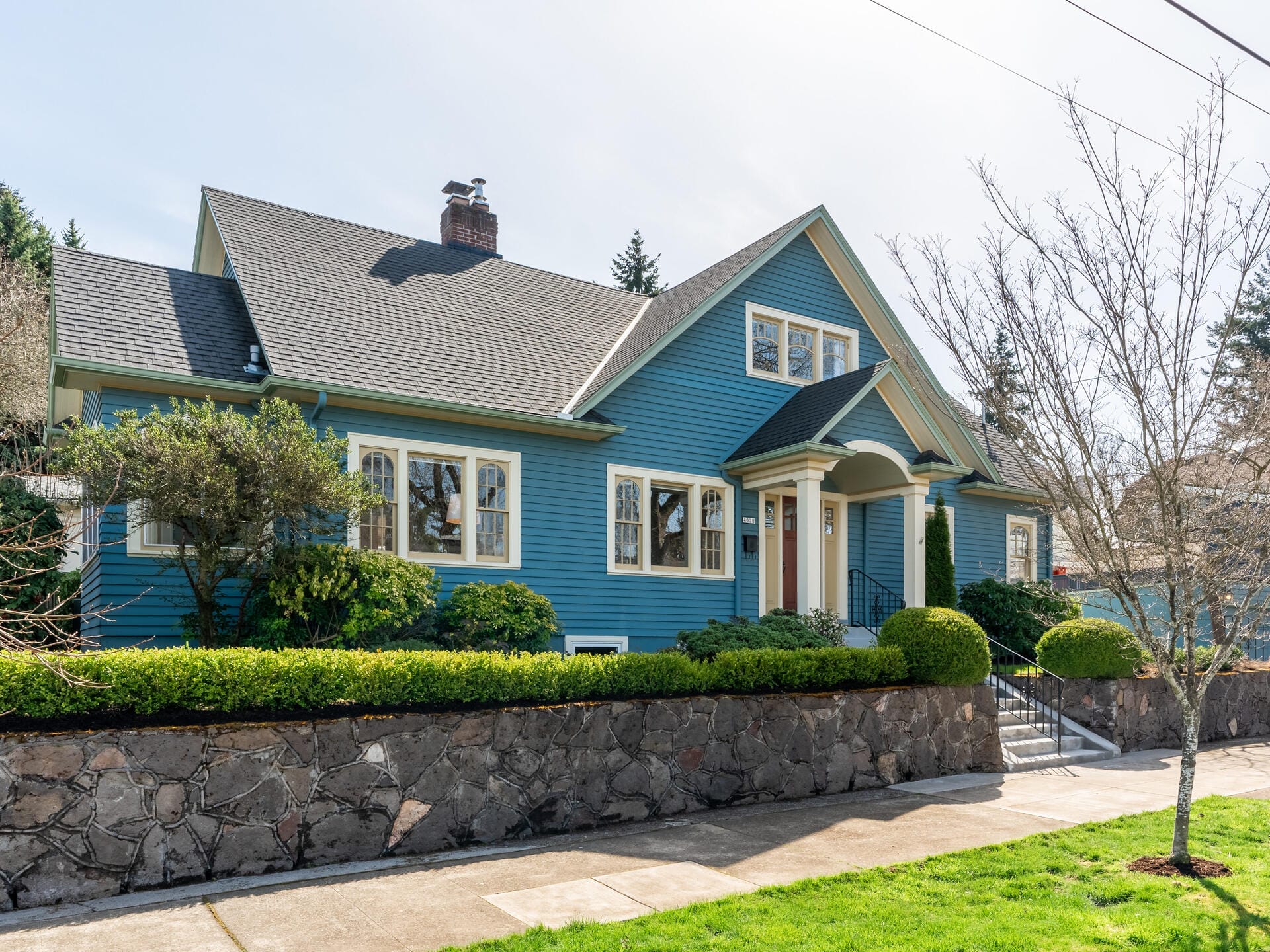 A charming blue house with white-trimmed windows and a steep, dark gray roof. It features a stone wall and lush greenery in the front yard, set against a clear sky. A sidewalk stretches along the front of the property.