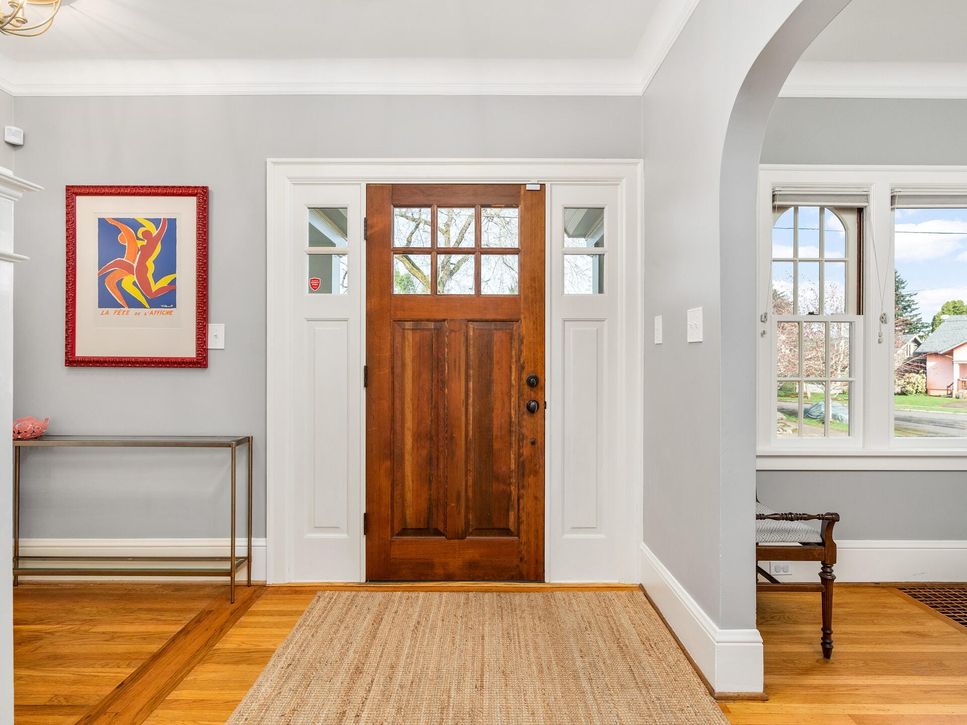 A welcoming home entrance with a wooden front door flanked by windows, standing on a light hardwood floor. A colorful framed artwork hangs on a gray wall, next to a sleek console table. Soft natural light floods the space through nearby windows.