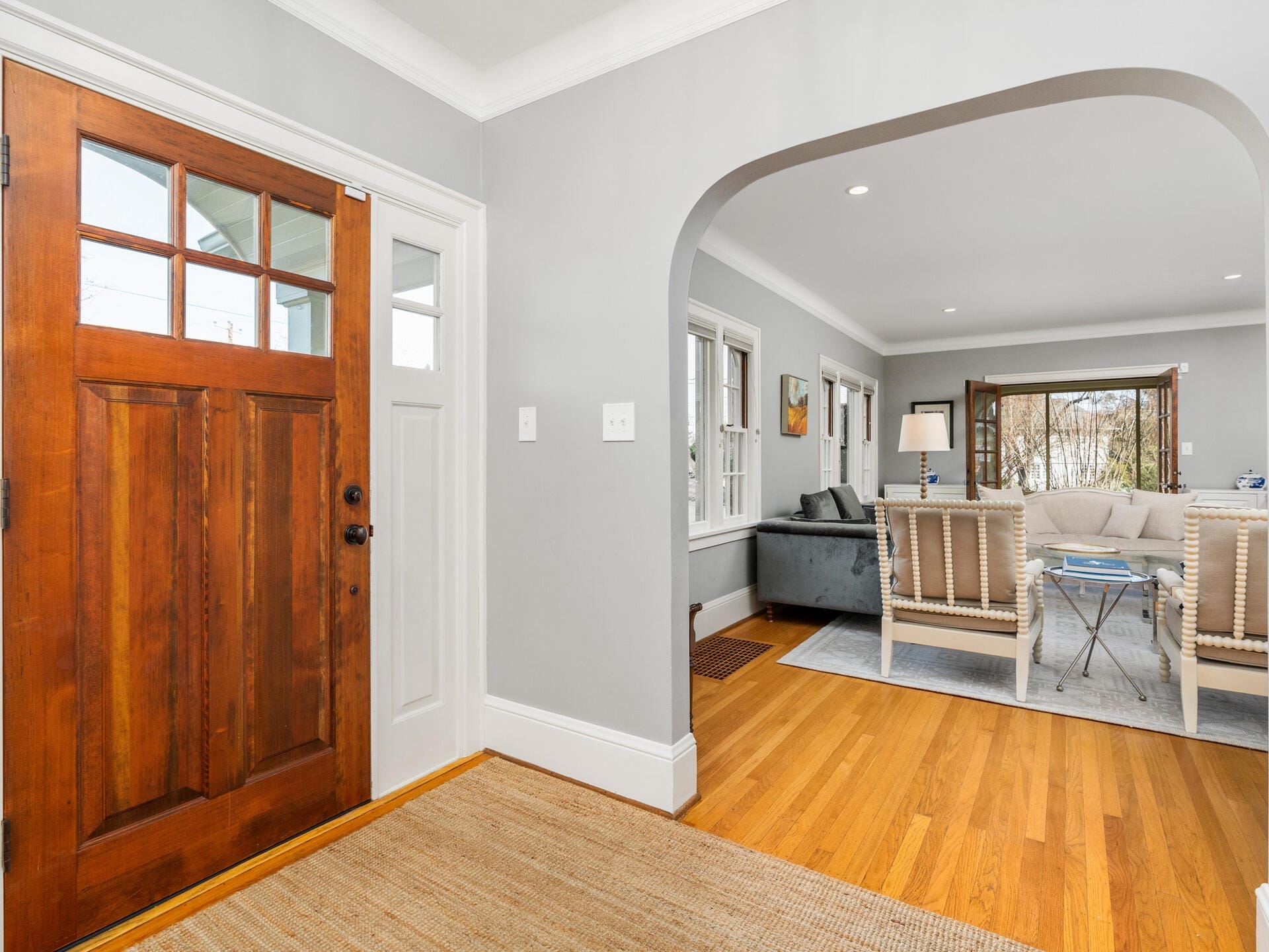 A cozy living room is visible through an arched doorway. The room features hardwood floors, a wooden front door on the left, a gray couch, two armchairs, a coffee table, and a large window allowing natural light to fill the space.
