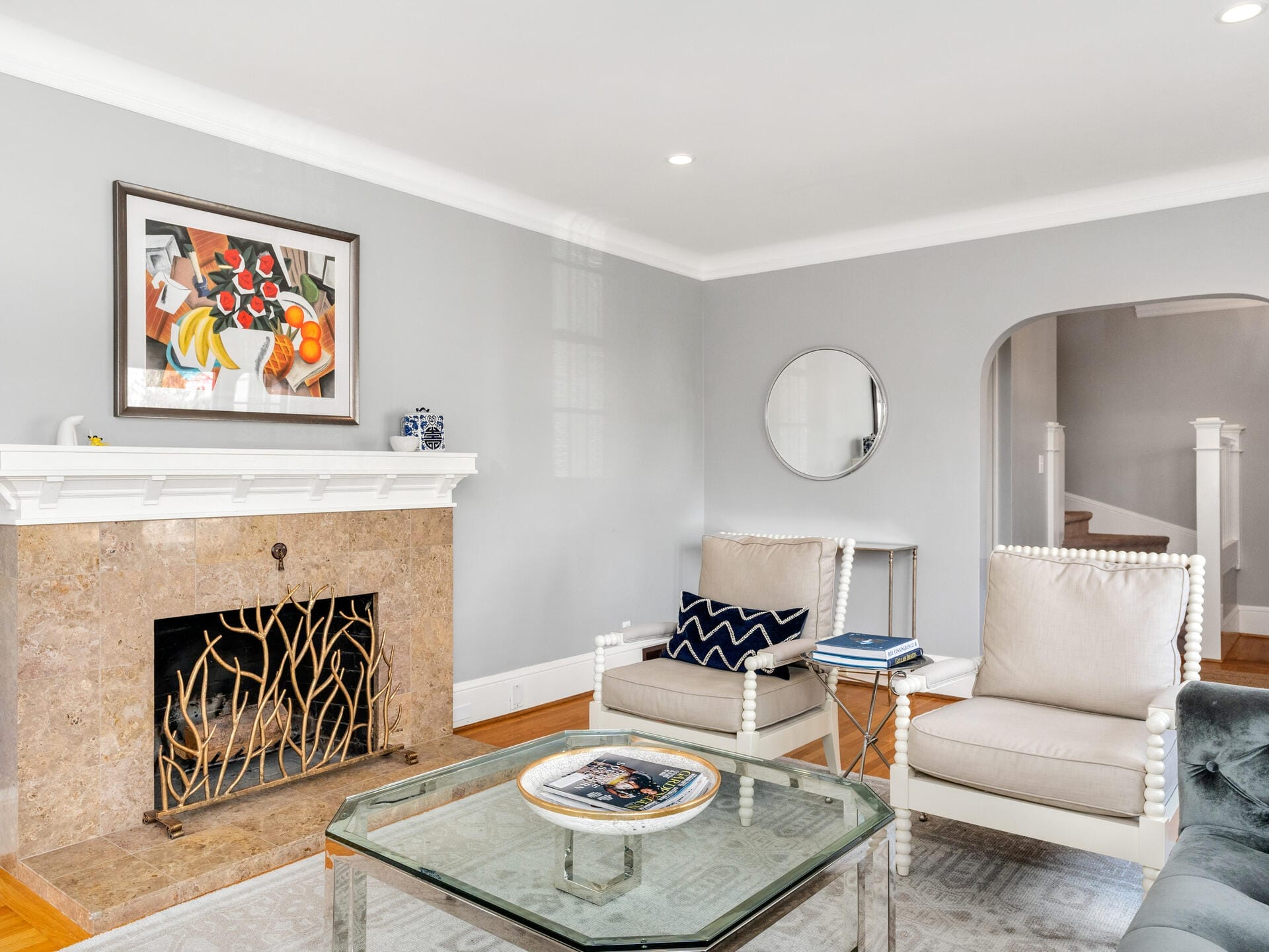 A modern living room with a light grey color scheme. Features include a marble fireplace with a decorative screen, two beige armchairs, a glass coffee table, a round wall mirror, and a colorful framed artwork above the mantel.