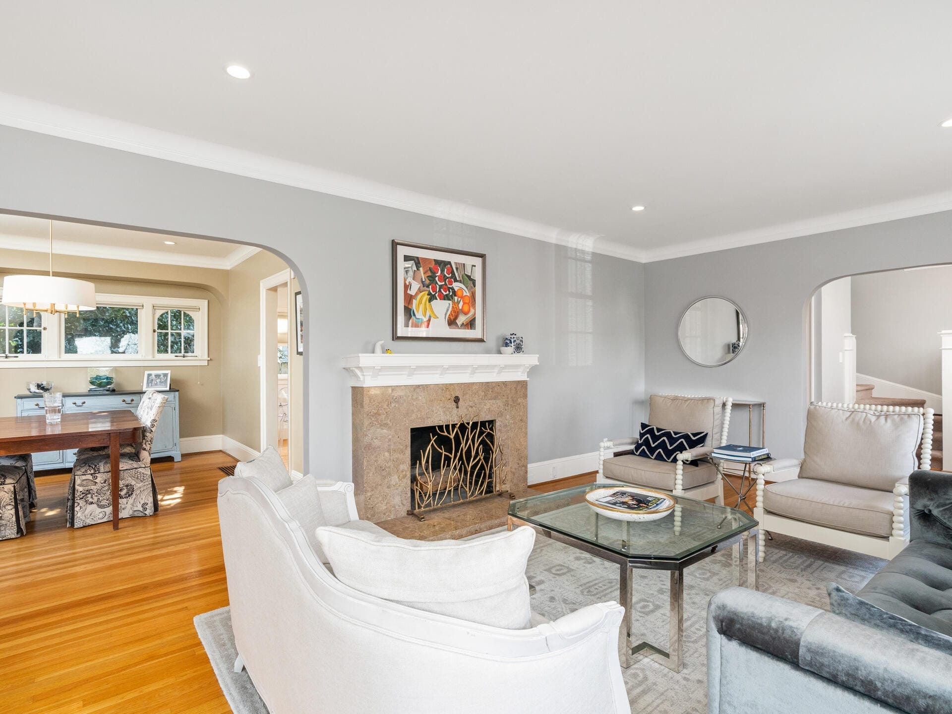A spacious living room with gray walls features a cozy sitting area, a fireplace with a painting above, and a glass coffee table. Adjacent is a dining room with a wooden table and cushioned chairs, seen through a wide archway.