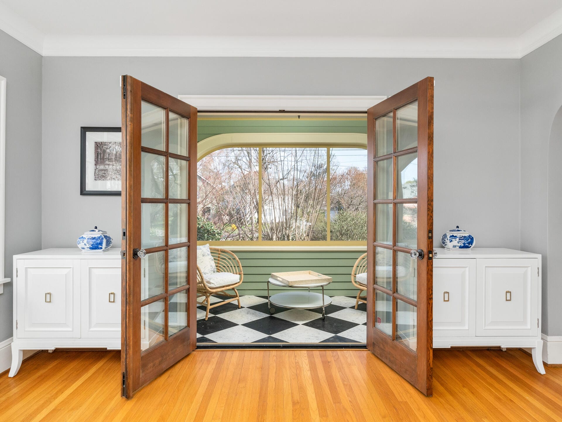A cozy sunroom with two wicker chairs and a small glass table is visible through open wooden French doors. The floor features a black and white checkered pattern, and there are white cabinets with blue and white vases on either side.