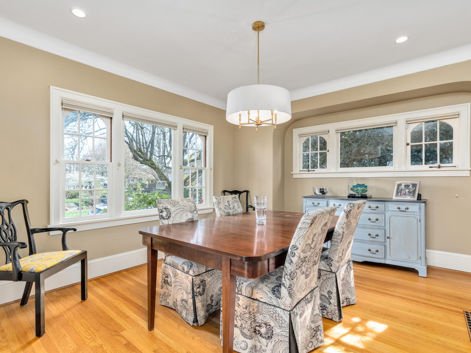 A cozy dining room with a wooden table and cushioned chairs. A large window offers views of trees outside. The room features hardwood flooring and beige walls. A white pendant light hangs above the table, and a blue sideboard holds framed photos.