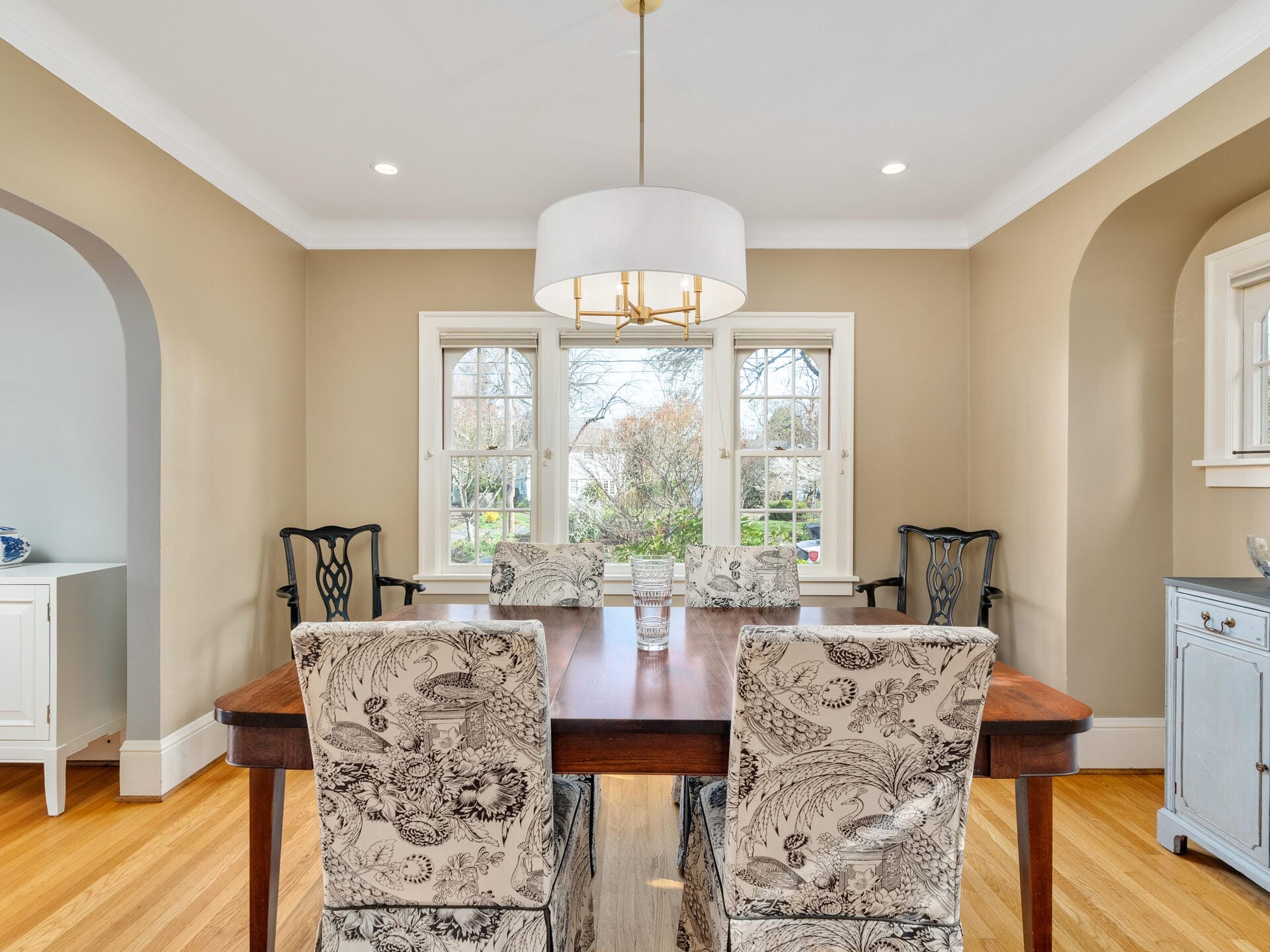 A dining room with a wooden table surrounded by six chairs featuring floral-patterned fabric. A round, white ceiling light hangs above the table. The space has beige walls, a hardwood floor, and windows offering a view of trees outside.