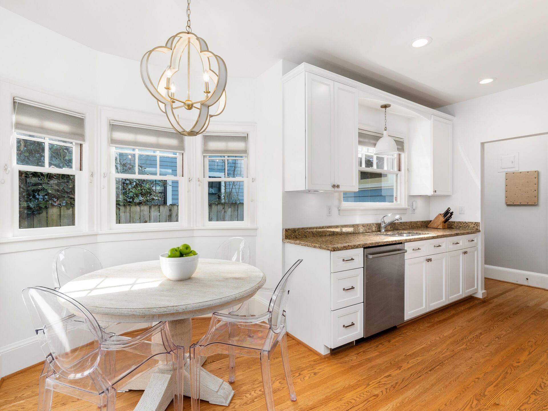 A modern kitchen with wooden flooring, white cabinets, and a granite countertop. A round dining table with clear chairs sits near three windows. A decorative chandelier hangs above the table, and a bowl of green apples is on the table.