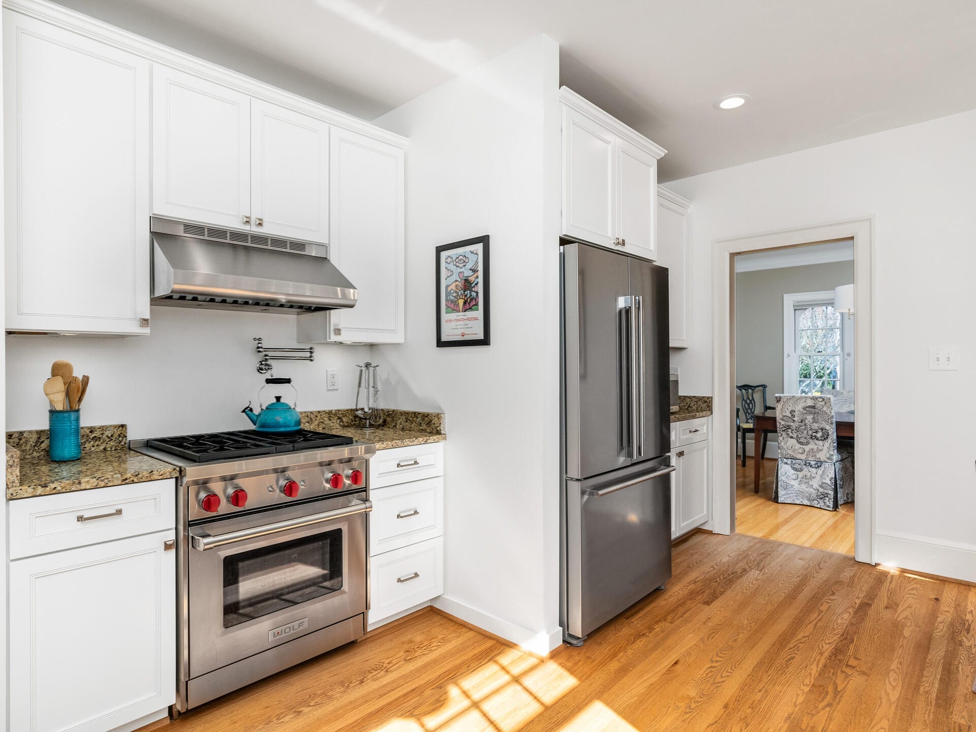 A modern kitchen featuring white cabinets, stainless steel appliances, and granite countertops. A blue teapot sits on the stove, utensils in a blue holder are on the counter, and a framed picture hangs on the wall. Wood flooring and open doorway visible.