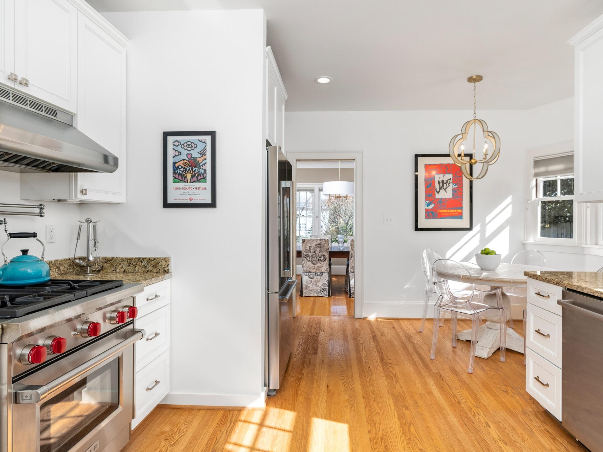 A bright kitchen with white cabinets and stainless steel appliances, including a range and dishwasher. Wooden floors lead to a dining area with a round table and clear chairs. Two colorful framed artworks decorate the walls. A window lets in natural light.