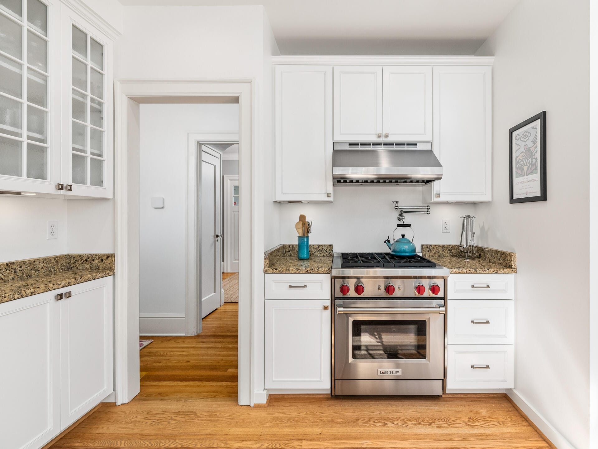 A modern kitchen with white cabinets and granite countertops. It features a stainless steel gas stove and oven, a range hood, and wood flooring. To the left are glass-front cabinets and an open doorway leading to a hallway.
