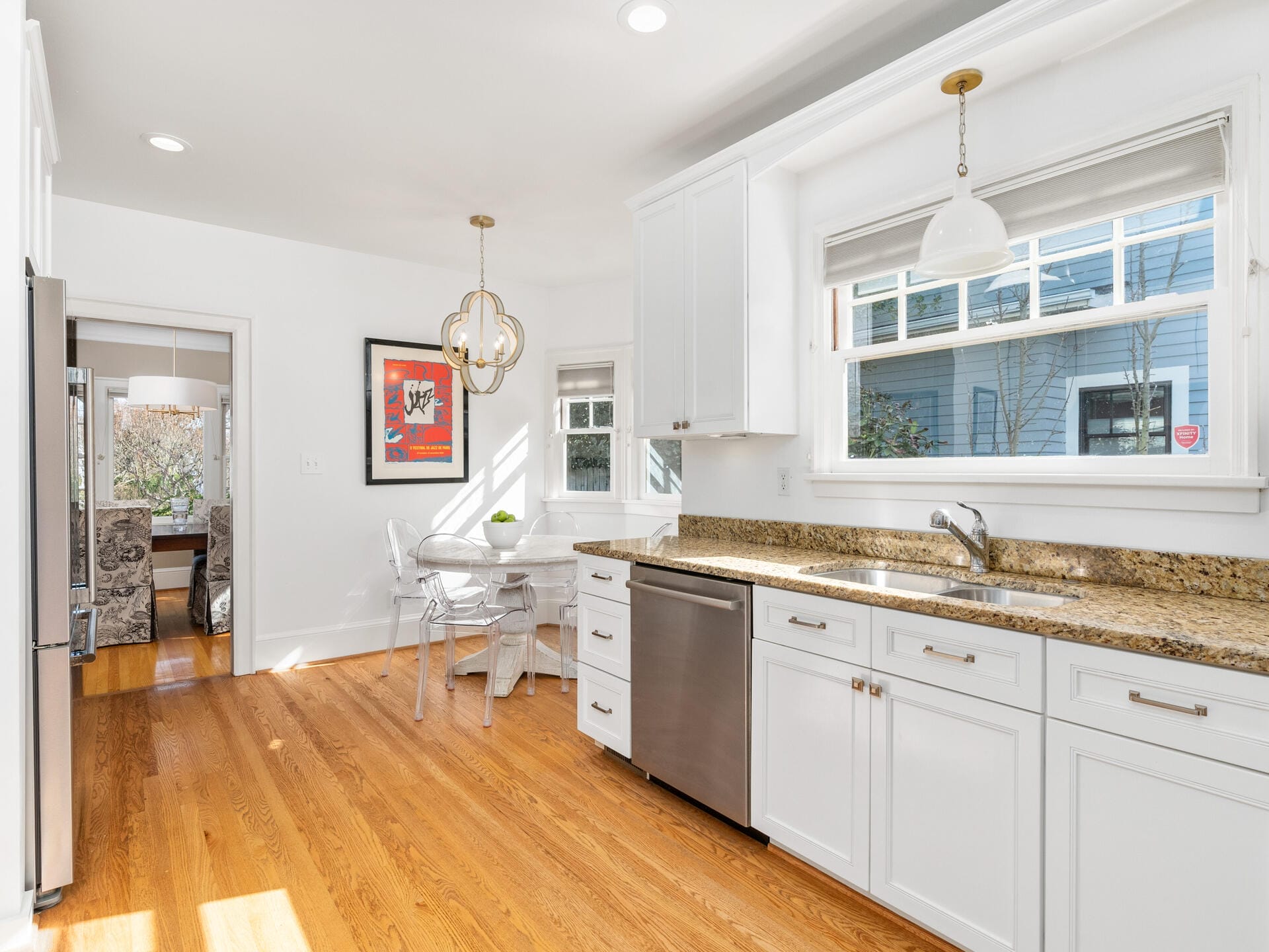 A bright kitchen with white cabinets, a dishwasher, and granite countertops. A window above the sink lets in natural light. A small dining area with a round table and modern chairs is near the window. Hardwood flooring throughout.