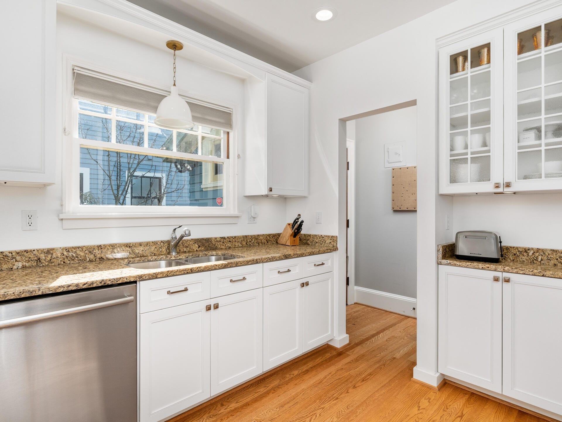 A bright kitchen with white cabinets and a window above the sink. The countertops are granite, and theres a stainless steel dishwasher. Hardwood floors complement the clean and modern look, with a toaster and knife block on the counter.