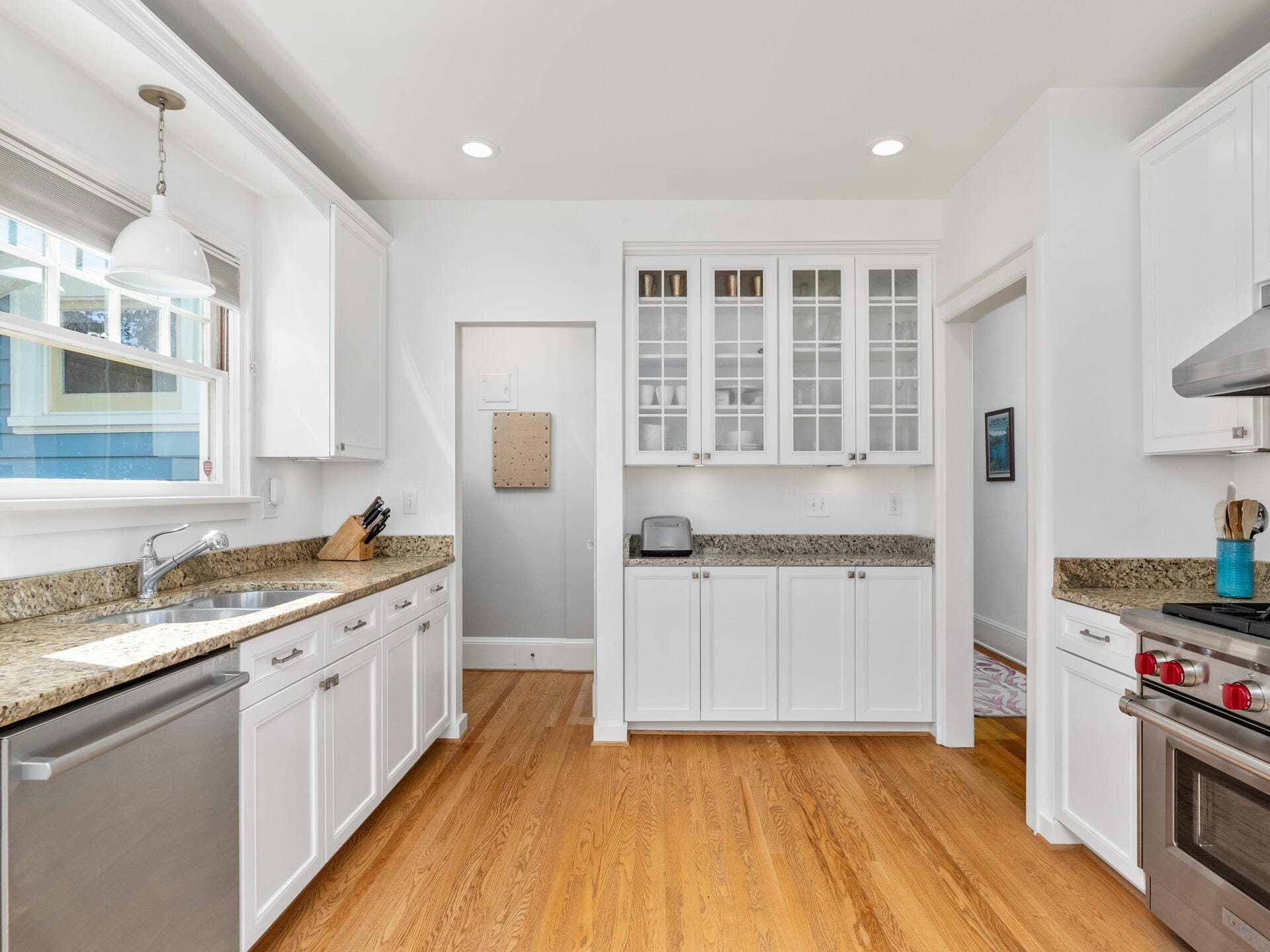 A modern kitchen with white cabinets, granite countertops, and stainless steel appliances. A large window lets in natural light. The room features a dishwasher, stove, and ample storage space. Light wood flooring complements the bright decor.