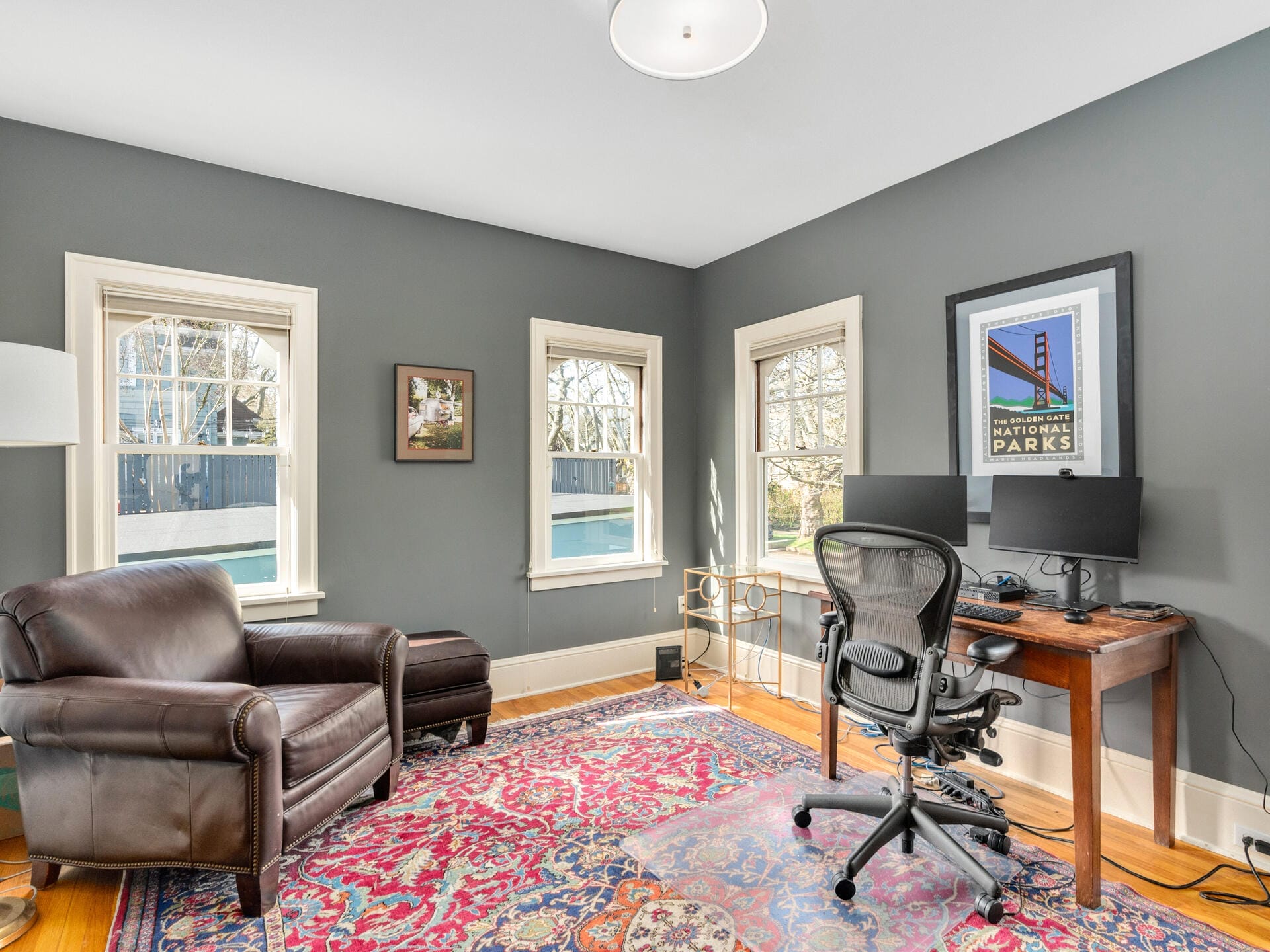 A cozy home office with gray walls features a large patterned rug, a leather armchair with an ottoman, and a wooden desk with dual monitors. Sunlight streams through three windows, casting light on a National Parks poster.