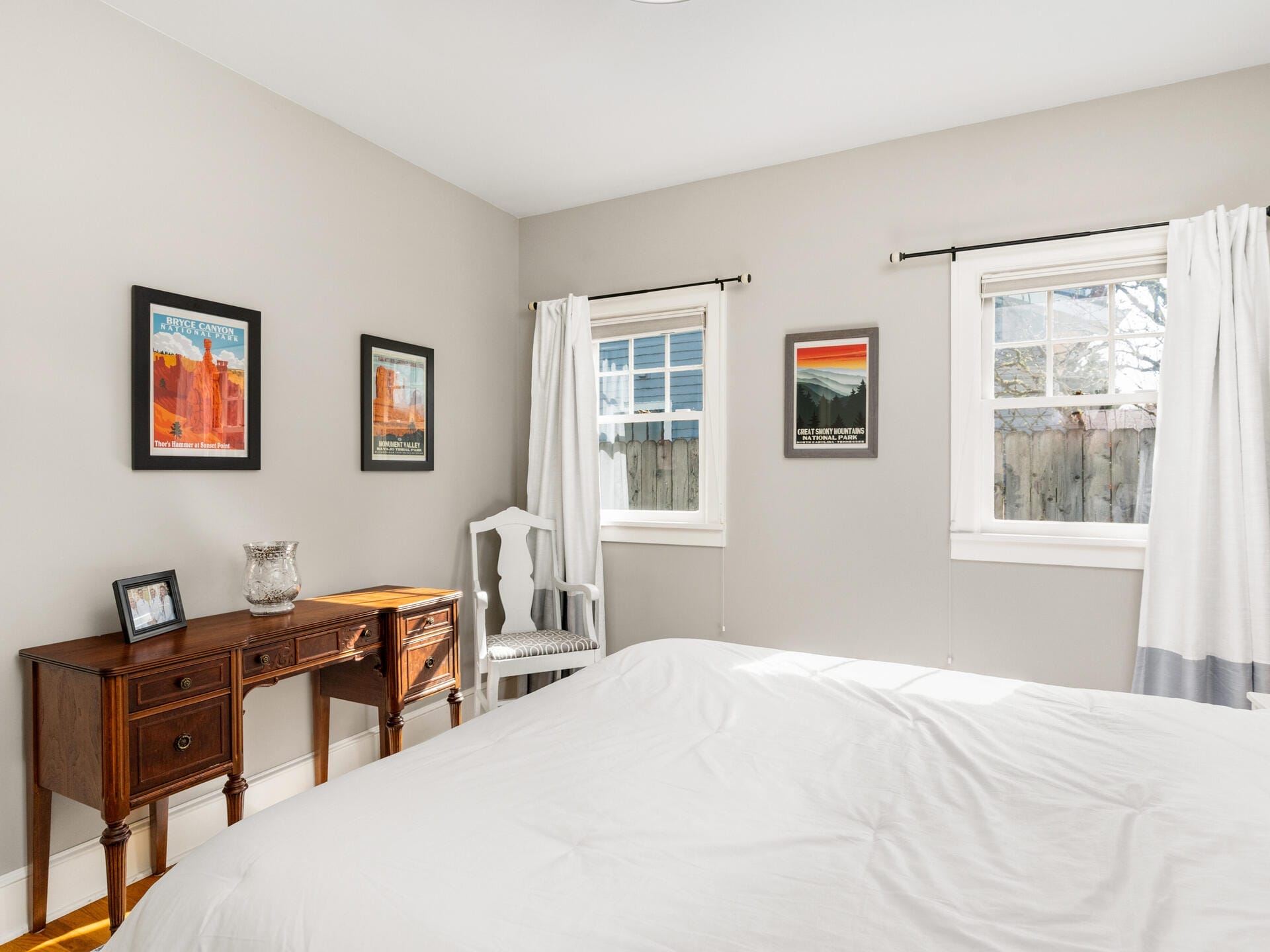 A bright bedroom with a neatly made bed, a wooden desk with a chair, framed pictures on light gray walls, and two windows with white curtains letting in natural light.