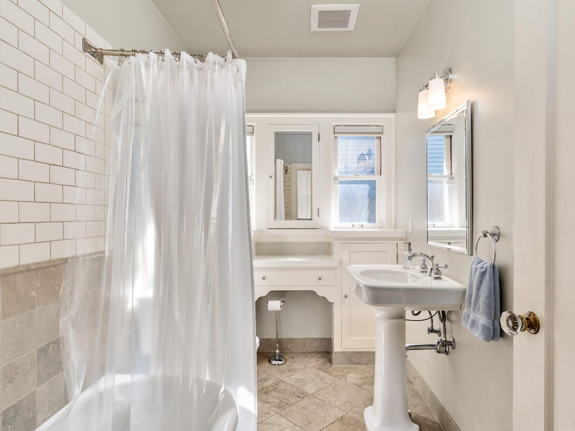 A bright bathroom with beige tiled floors and white subway tile walls. It features a pedestal sink, a mirror, a towel holder with a blue towel, a bathtub with a shower curtain, a window, and a wall-mounted cabinet.