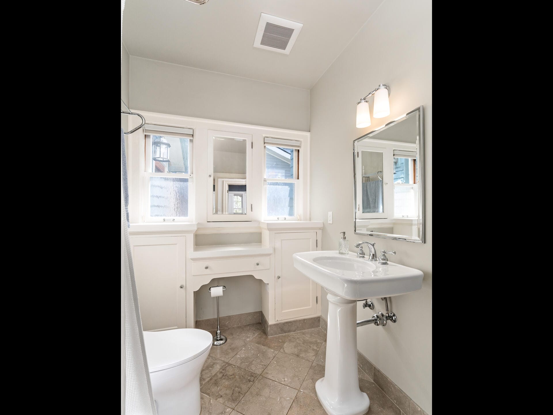A bright bathroom features a pedestal sink with a mirror above, a toilet, and a window with white trim. Theres also a built-in vanity under the window. The walls and floor are light-colored, enhancing the spacious feel.