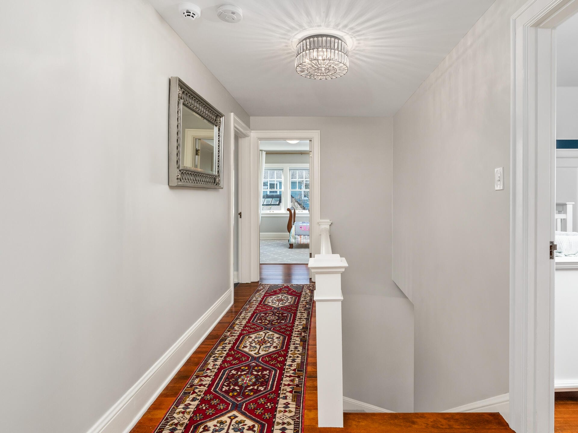 A hallway with a wooden floor features a patterned red runner rug. A decorative mirror hangs on the left wall. At the end of the hallway, a room with a bed is visible. A chandelier-style light fixture is on the ceiling.