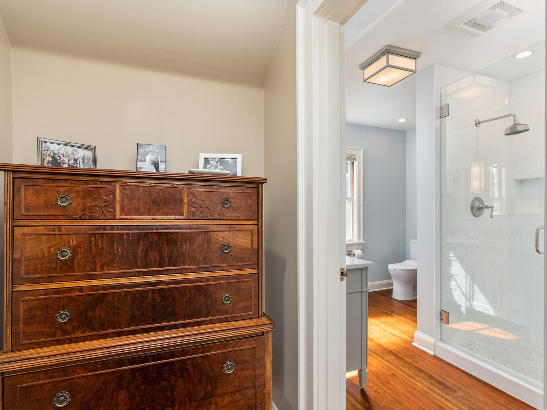 A cozy room featuring a vintage wooden dresser with framed photos on top. Adjacent is a modern bathroom with a glass shower, natural light streaming through a window onto the wooden floor.