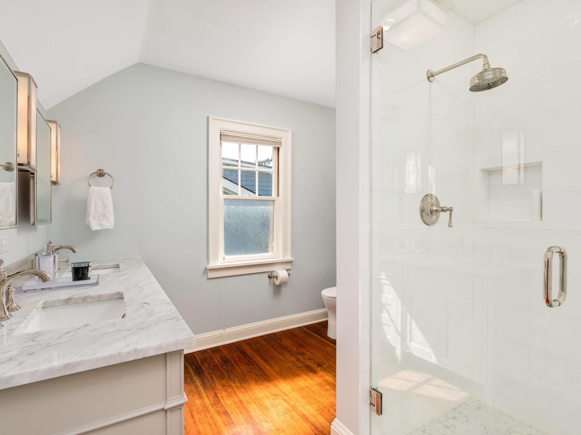 A bright bathroom with wooden floors, a white dual-sink vanity, a towel hanging on the wall, and a window above. To the right is a glass-enclosed shower with a rainfall showerhead.