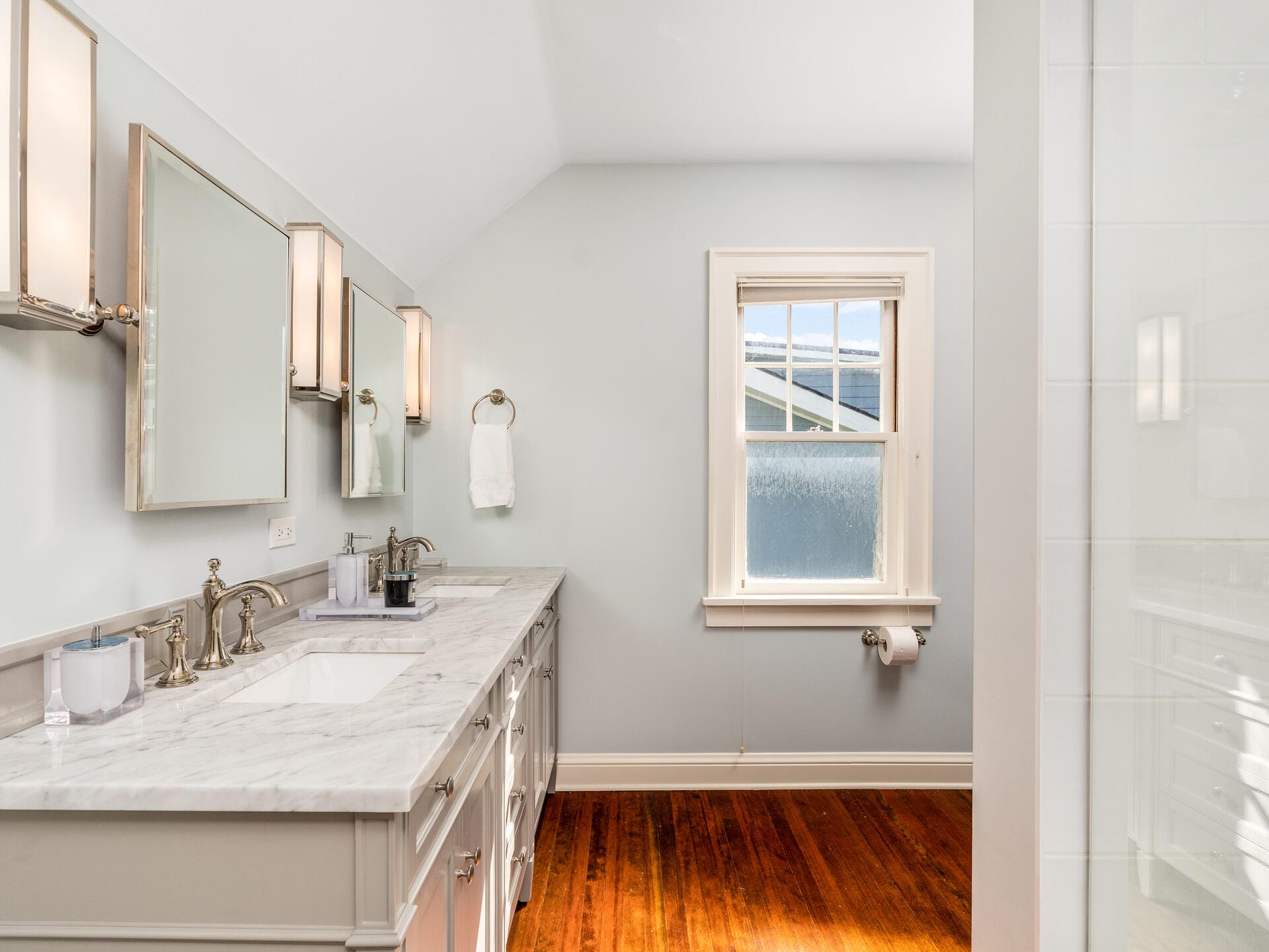 A bright bathroom features a double sink vanity with marble countertops, large mirrors, and elegant lighting. There is a window with frosted glass, wood flooring, and neatly folded towels by the sinks.