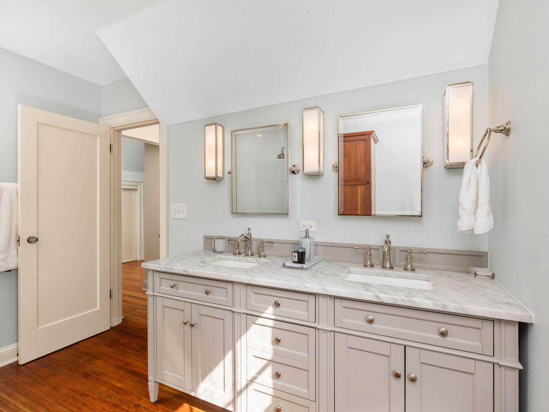 A modern bathroom features a double sink vanity with a marble countertop, two mirrors, and wall-mounted light fixtures. The room has hardwood floors, light-colored walls, and an open door leading to an adjoining area.